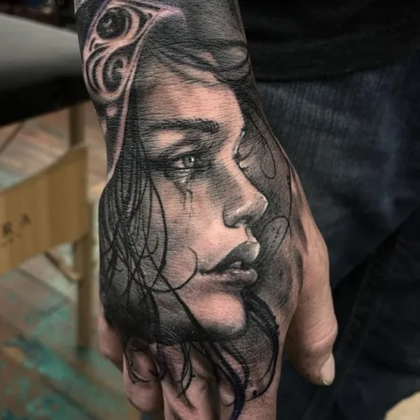 Black and gray realism tattoo on the hand of a woman's crying profile with a tribal eye design