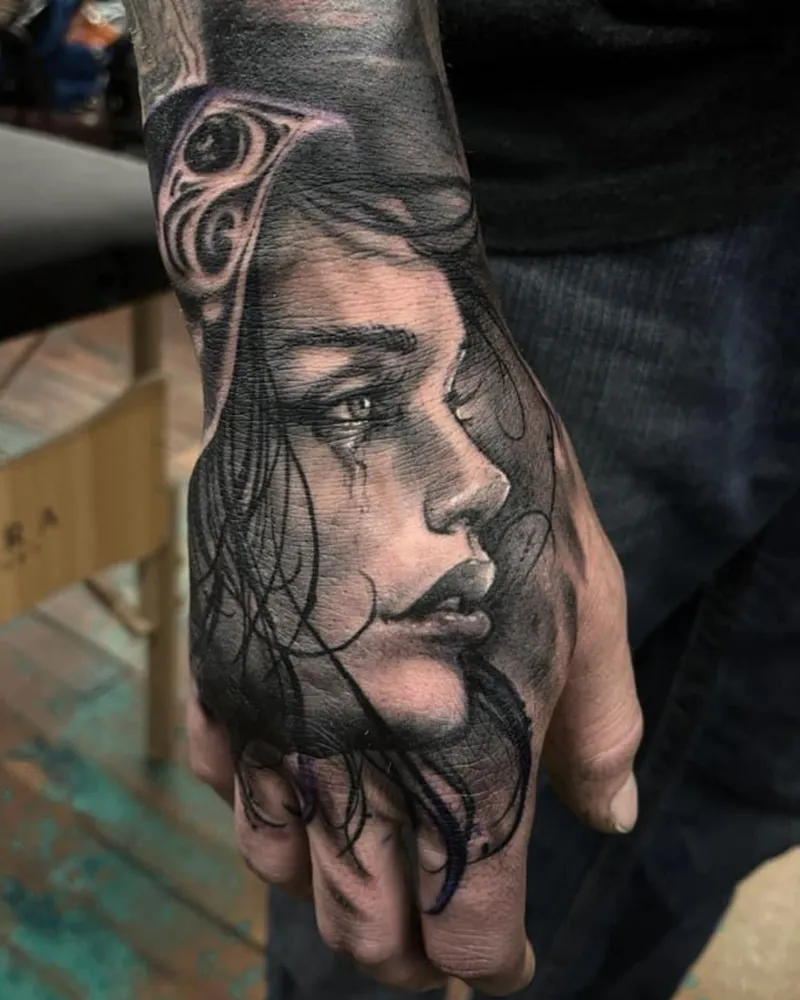 Black and gray realism tattoo on the hand of a woman's crying profile with a tribal eye design