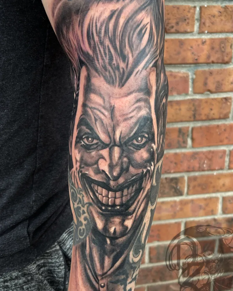 Black and gray tattoo of a sinister, grinning joker-like face on the forearm