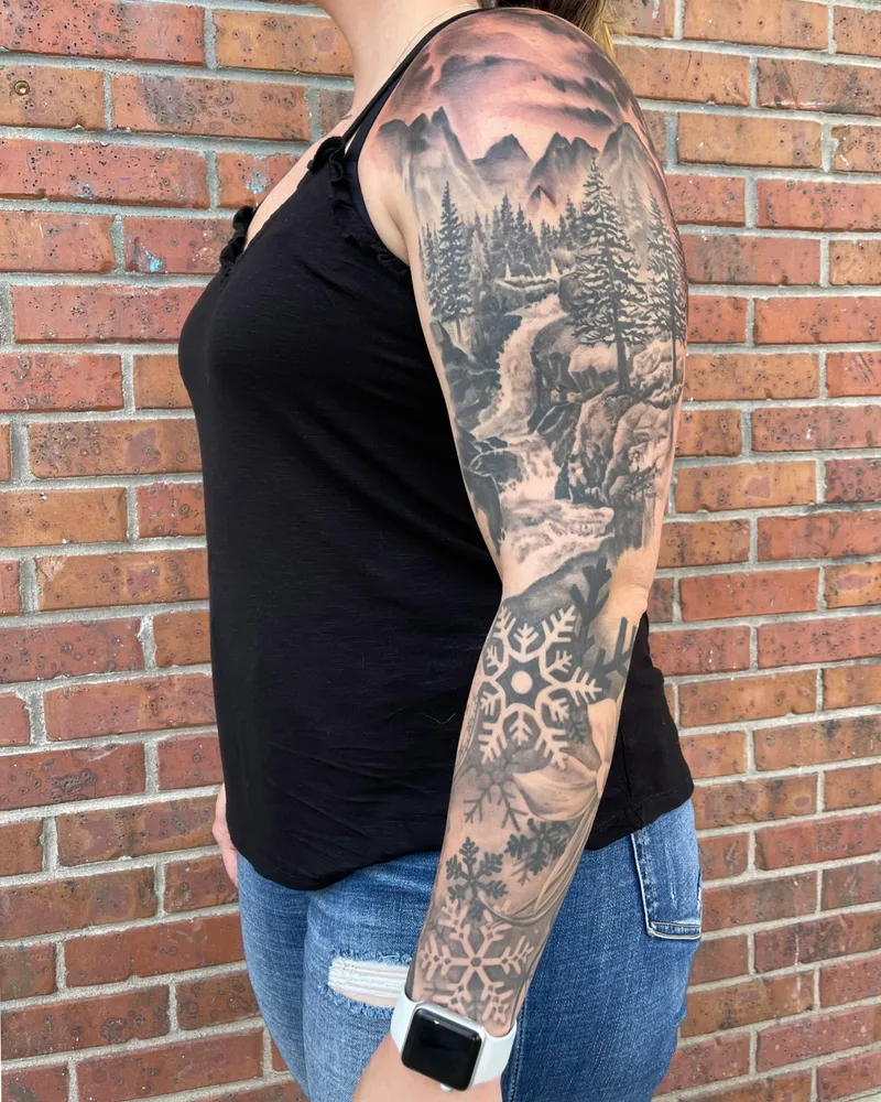 Black and gray full sleeve of a mountain waterfall landscape with pine trees, flowing into snowflakes down the forearm