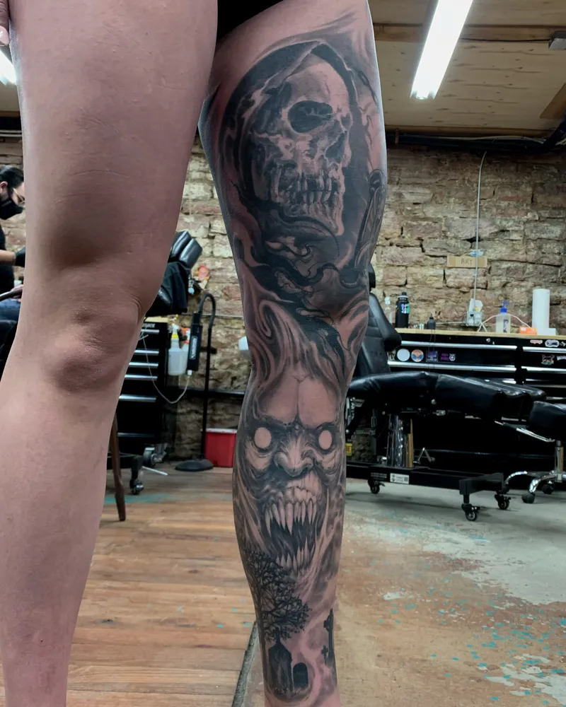 Full leg sleeve with a hooded reaper skull and a fanged demon face above a graveyard at the ankle