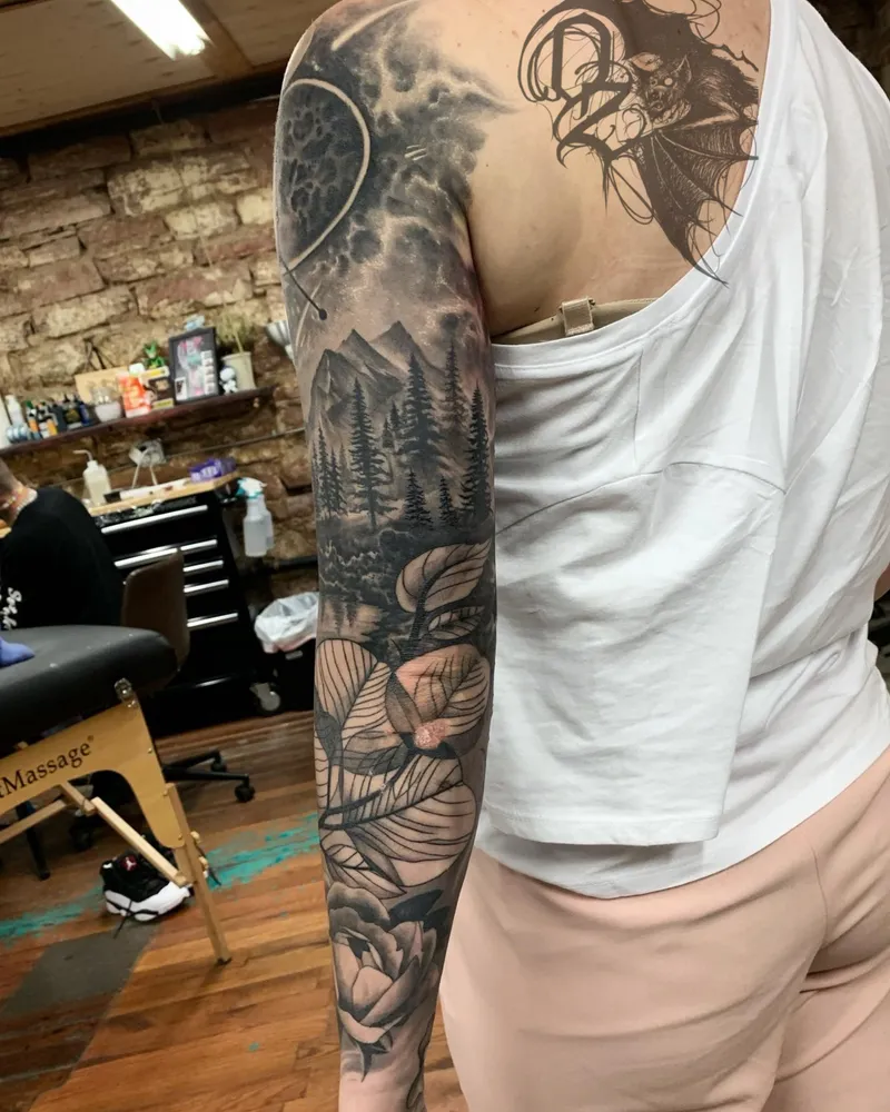 Back view of a full sleeve with a moon over a mountain forest, finishing in linework leaves and a rose