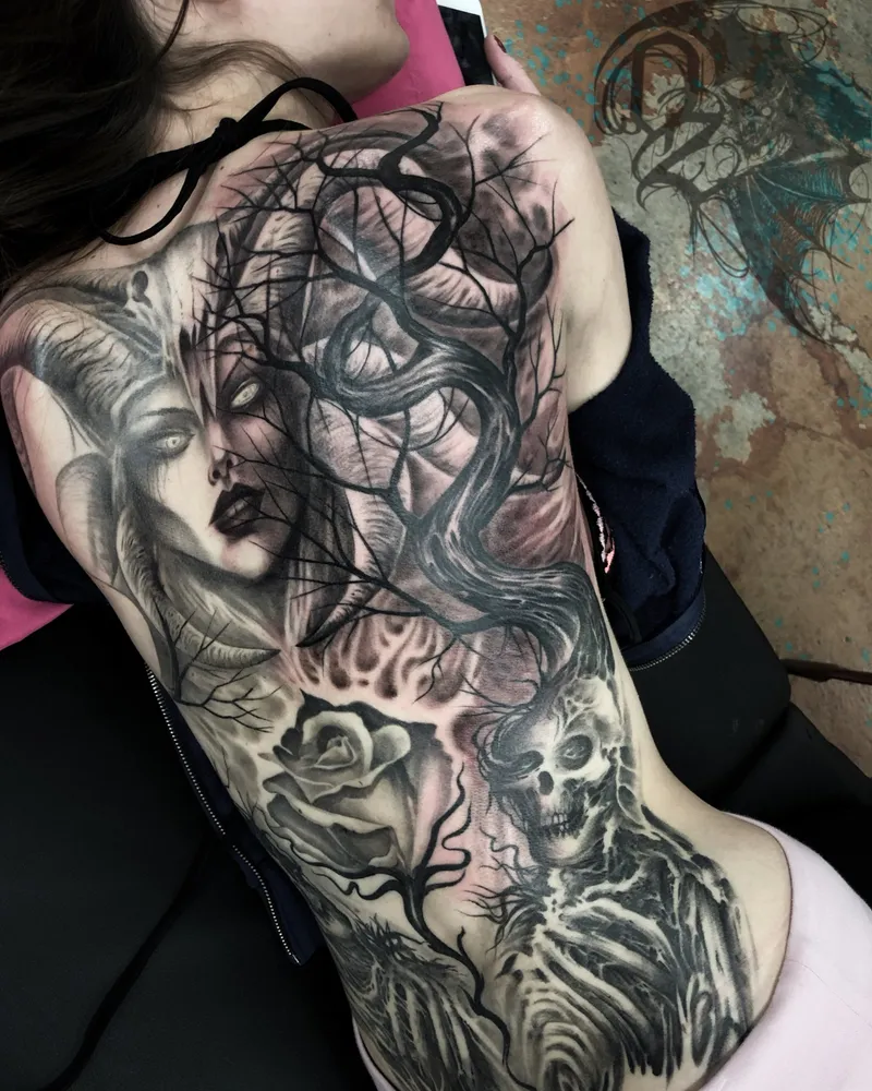 Full back piece of a horned woman among bare branches, with a rose and a skeletal figure on the lower back