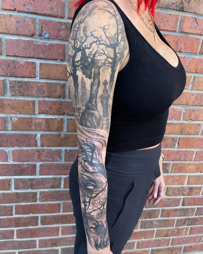 Full sleeve with a cemetery angel under a full moon and a woman's eye emerging from bare branches on the forearm